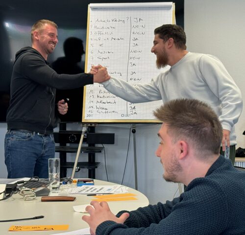 Zwei Teilnehmer begrüßen sich per Handschlag vor einem Flipchart während eines Grossmann-Training-Seminars